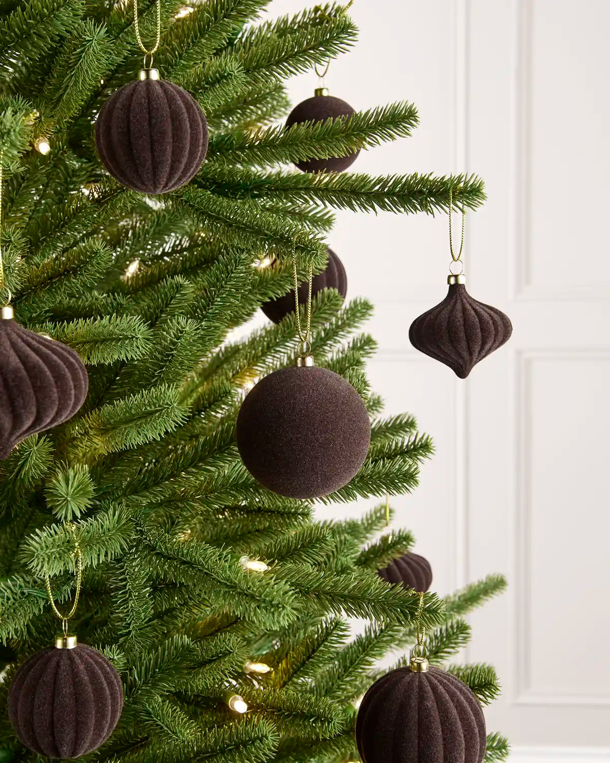 Dark brown velvet Christmas tree ornaments hanging on a green pine tree with warm white string lights, set against a white paneled wall.