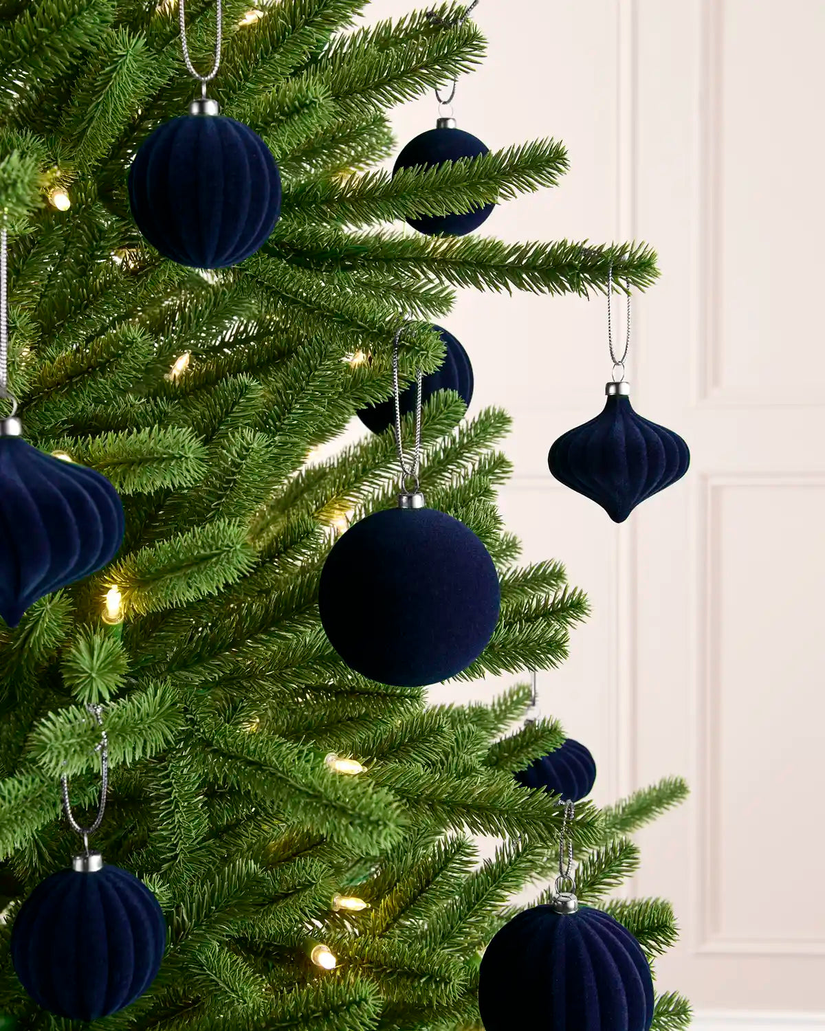 Dark navy blue velvet Christmas tree ornaments hanging on a green artificial Christmas tree with warm white string lights, set against a light cream paneled wall.