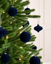 Dark navy blue velvet Christmas tree ornaments hanging on a green artificial Christmas tree with warm white string lights, set against a light cream paneled wall.