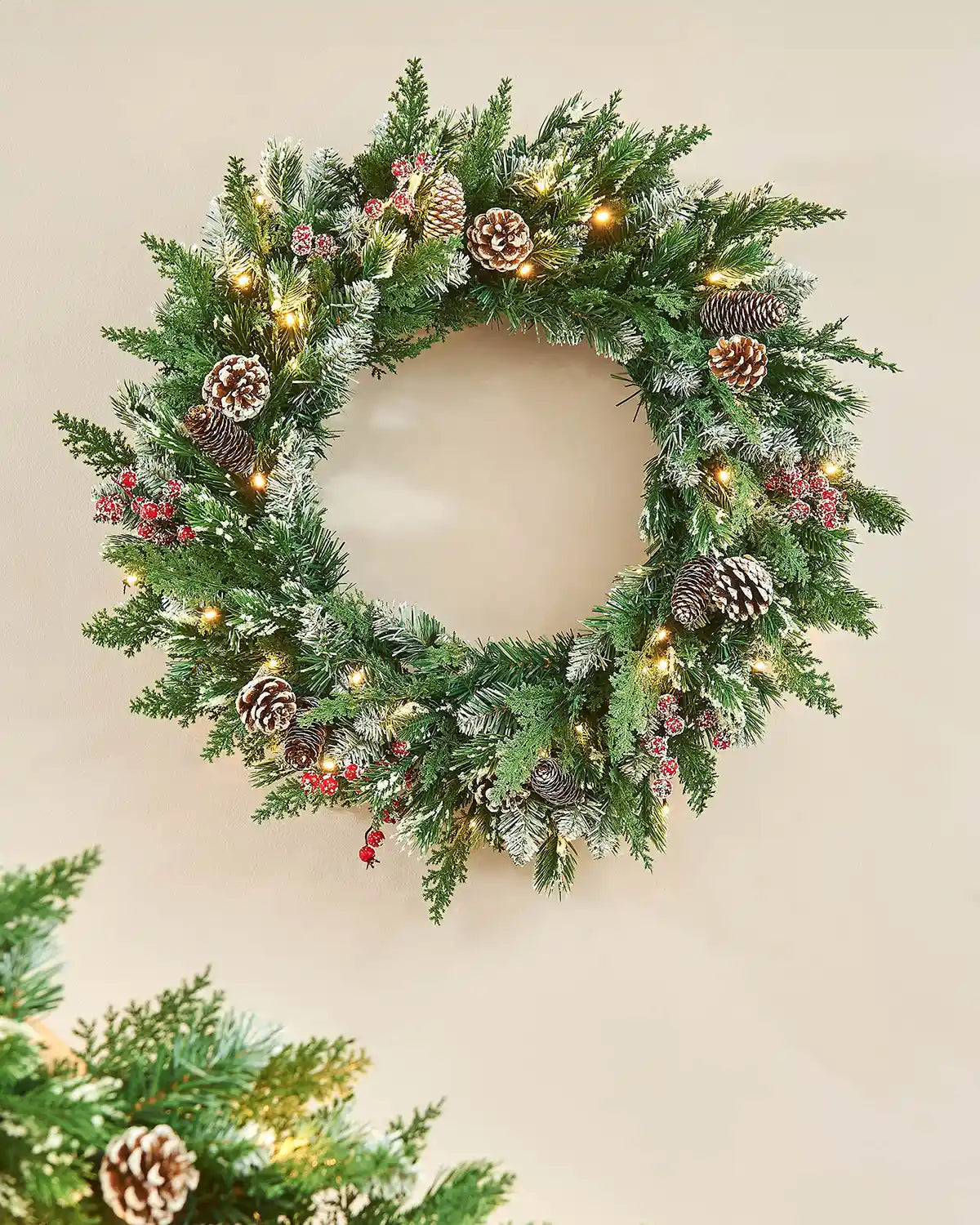 Decorated Christmas frosted wreath with pinecones, berries and lights on a cream wall 
