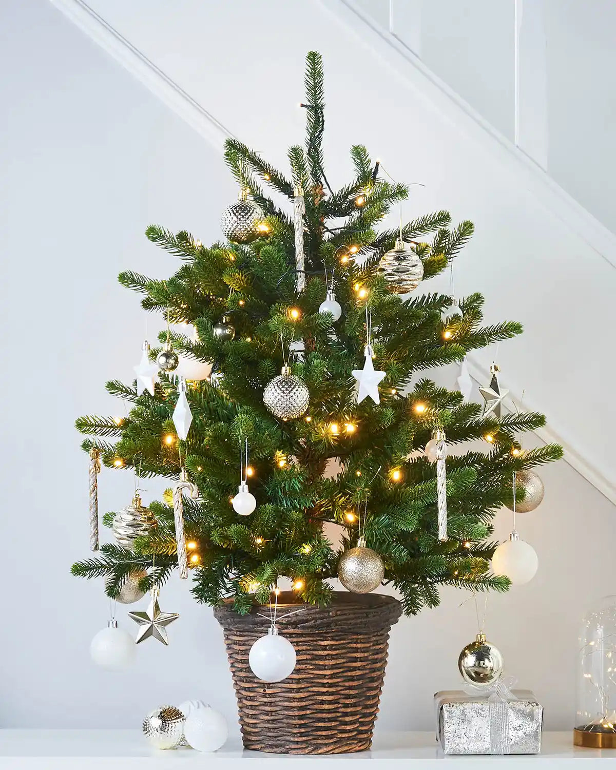 Decorated Christmas tree with lights and ornaments in a basket on a white surface in front of staircase 
