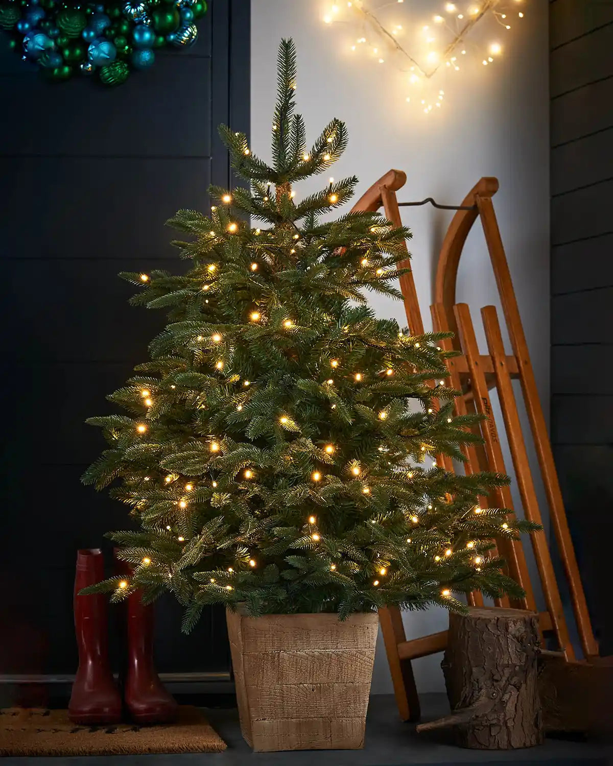 Decorated Christmas tree with lights in a textured square wooden pot on a doorstep with a wooden sled and wellington boots 