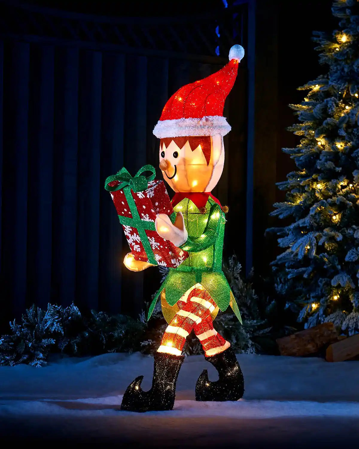 Decorative Christmas elf holding a gift in front of a lit Christmas tree in the garden