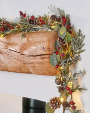 Decorative Christmas garland with Red berried, pinecones with lights on a wooden mantel against a white wall.