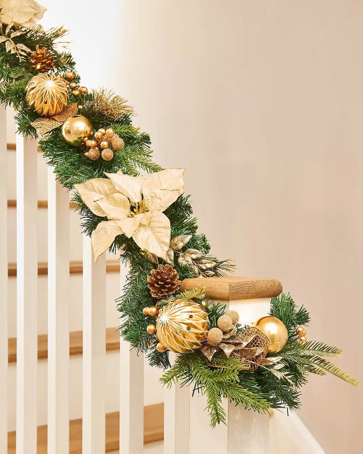 Decorative Christmas garland with gold poinsettias, pinecones, and lights on a staircase banister against a cream background.