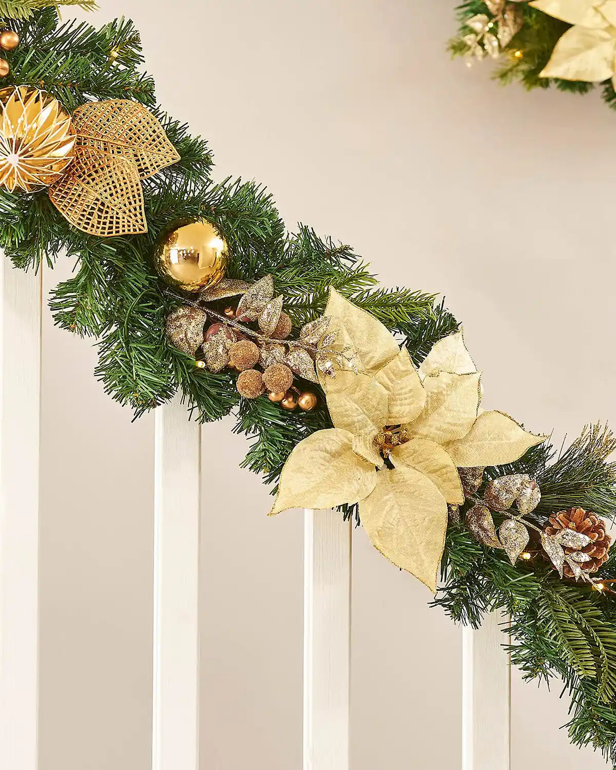 Decorative Christmas garland with gold poinsettias, pinecones, and lights on a staircase banister neutral background.