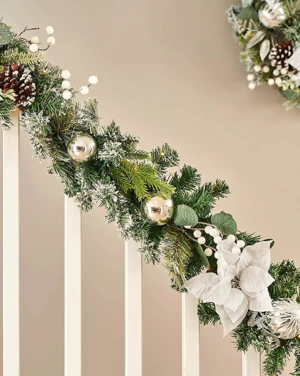 Decorative Christmas garland with greenery, ornaments, and a white bow on a wooden staircase banister against a beige wall