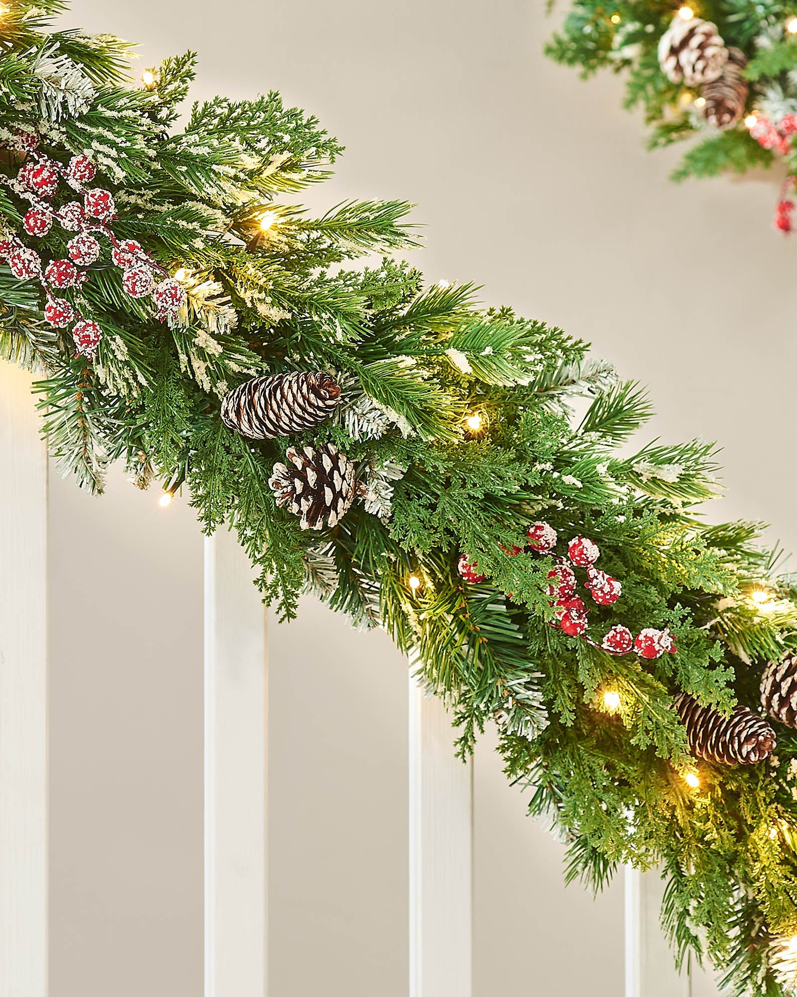 Decorative Christmas garland with lights, pine cones, and berries on a  staircase banister neutral wall.