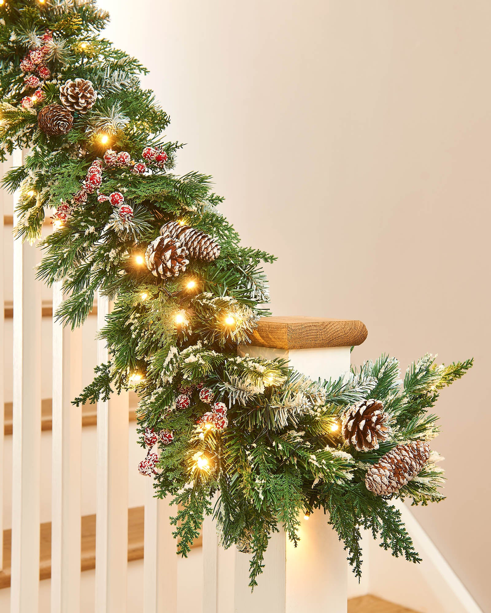 Decorative Christmas garland with lights, pine cones, and berries on a wooden staircase banister neutral wall.