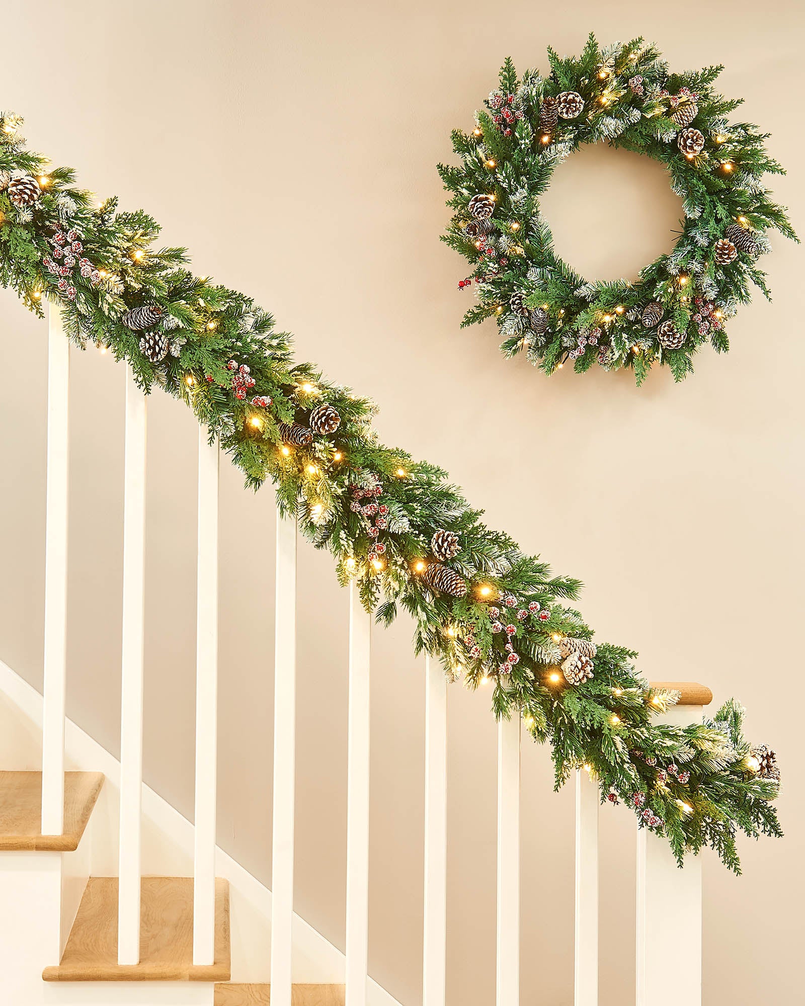 Decorative Christmas garland with lights, pine cones, and berries with a matching wreath on a staircase banister neutral wall.
