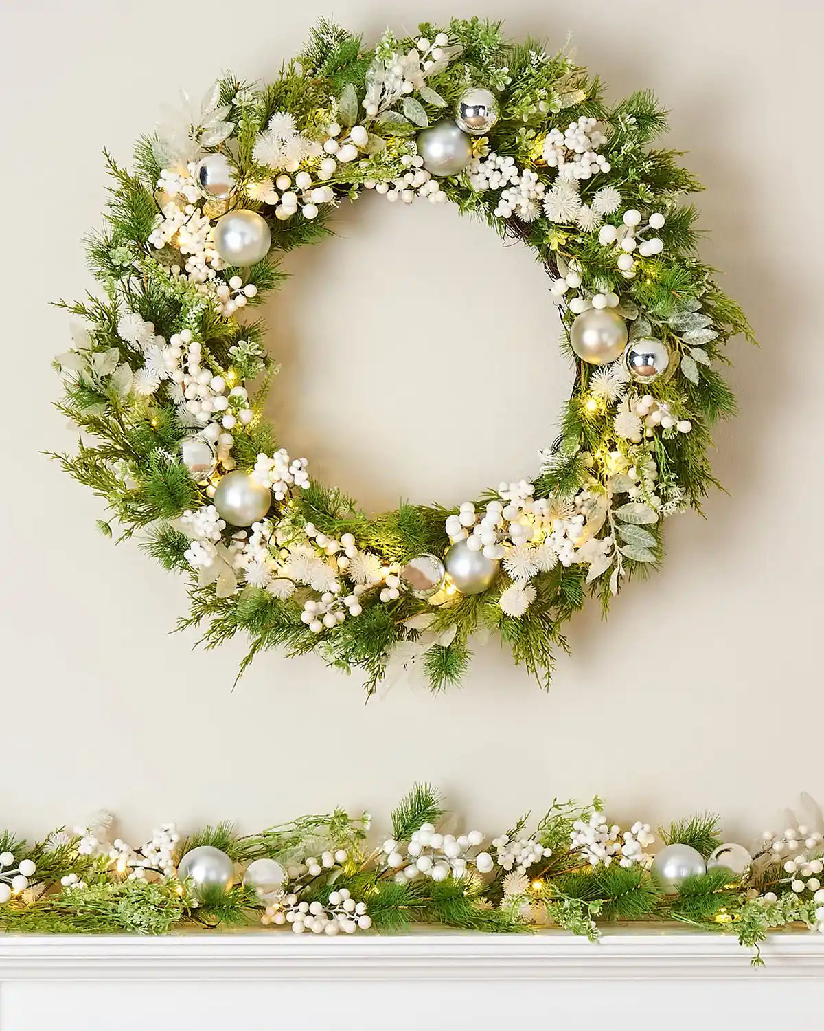 Decorative Christmas wreath with greenery, white flowers, and silver baubles with matching garland on a white background
