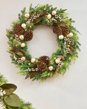Decorative Christmas wreath with pine cones, berries, and lights on a cream wall.