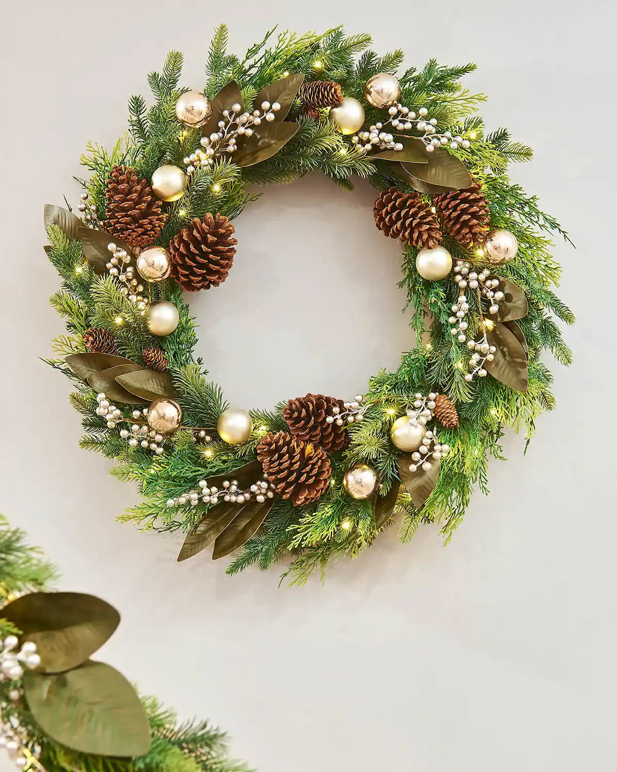 Decorative Christmas wreath with pine cones, berries, and lights on a cream wall.