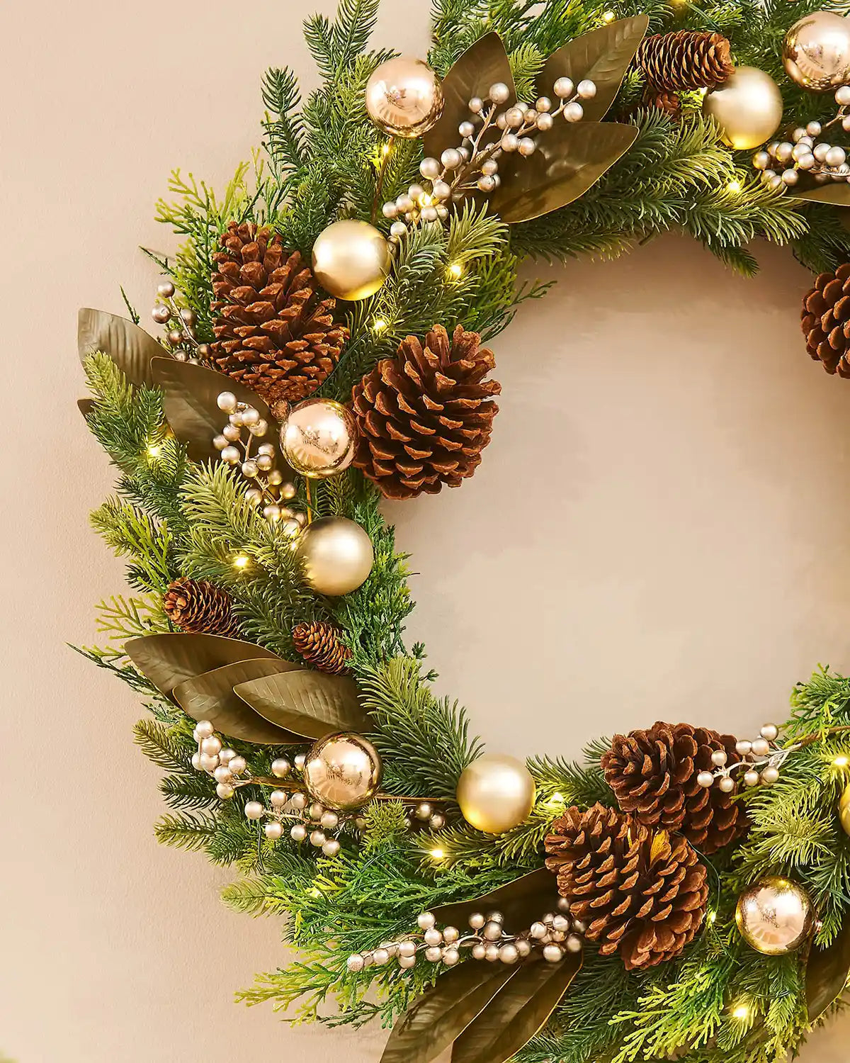Decorative Christmas wreath with pine cones, gold baubles, and lights on a beige wall.