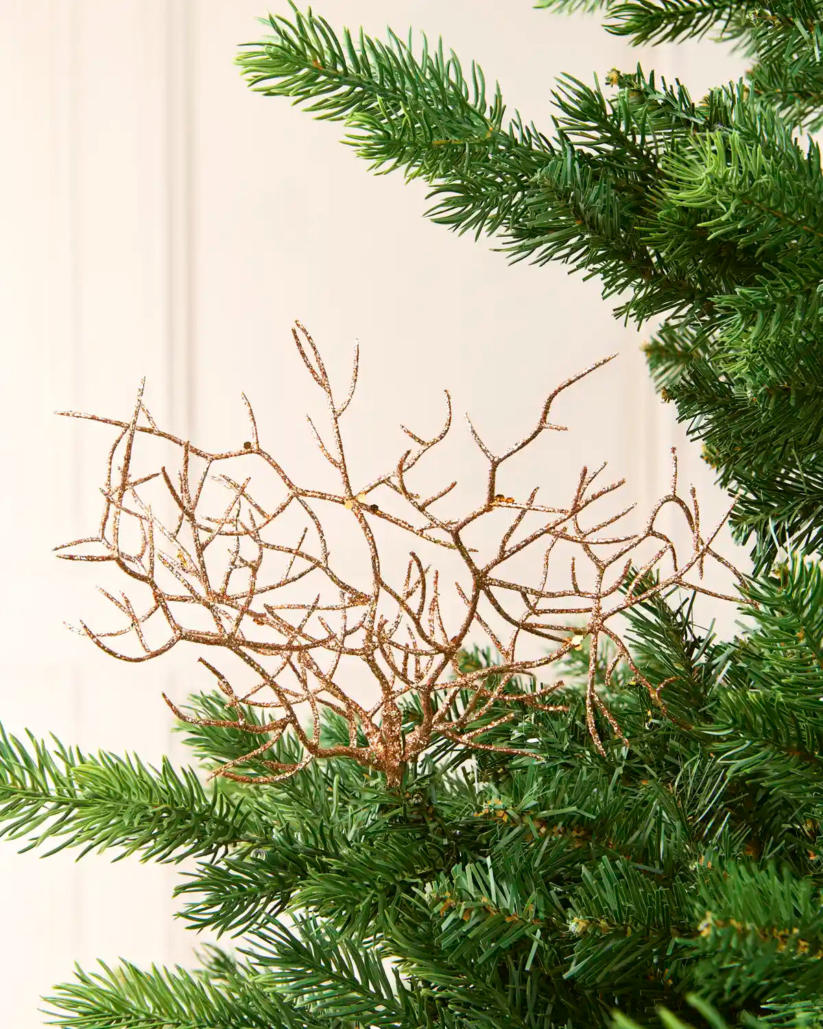 Decorative Rose-gold glitter branch ornament with a textured, twig-like structure, shown against a green Christmas tree needles and a light cream wall.