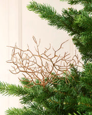 Decorative Rose-gold glitter branch ornament with a textured, twig-like structure, shown against a green Christmas tree needles and a light cream wall.