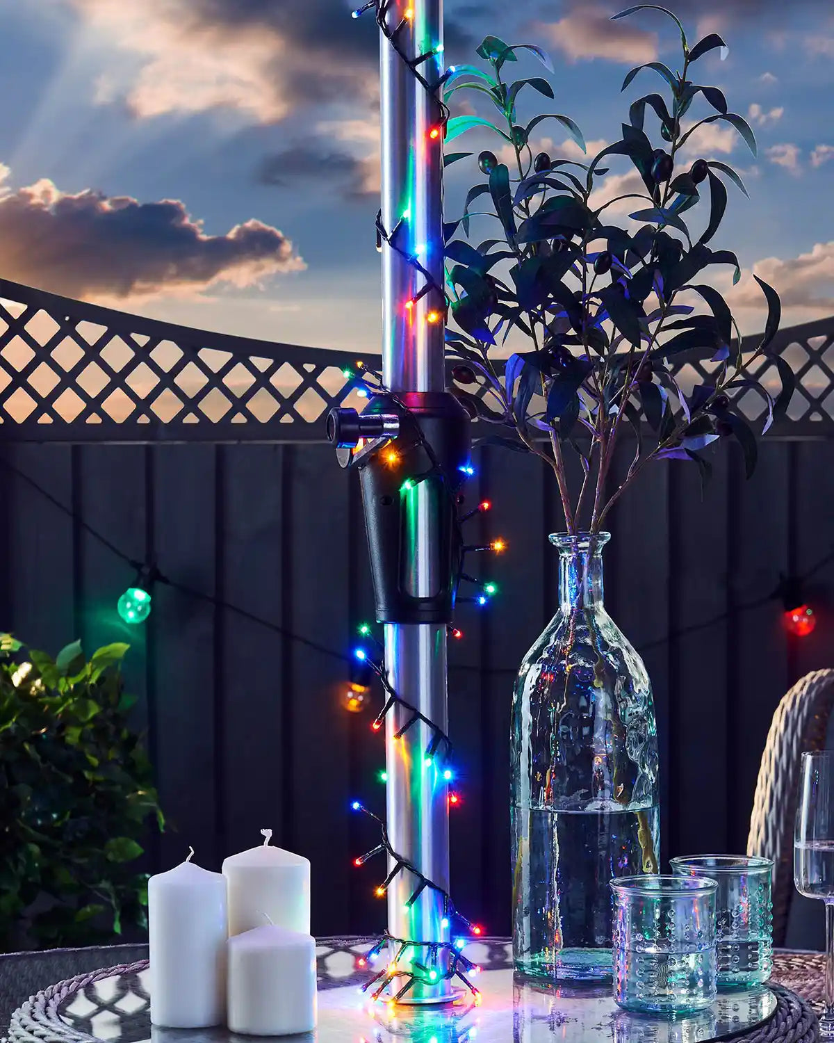 Decorative garden setup with bottles, candles, multi coloured lights on umbrella post and plants against a sunset sky.