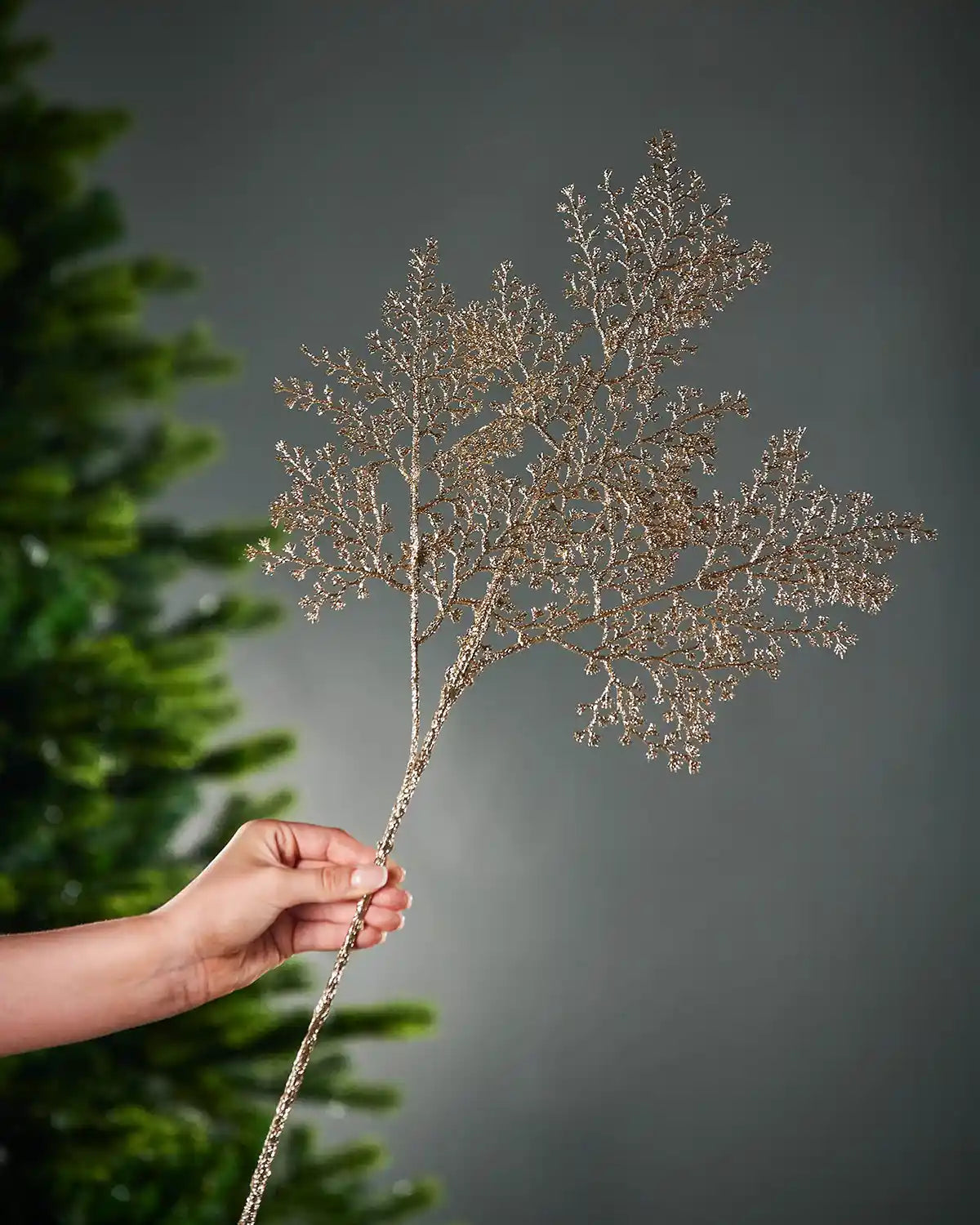 Decorative gold glitter artificial plant stem with small branches held by a hand with a Christmas tree in the background