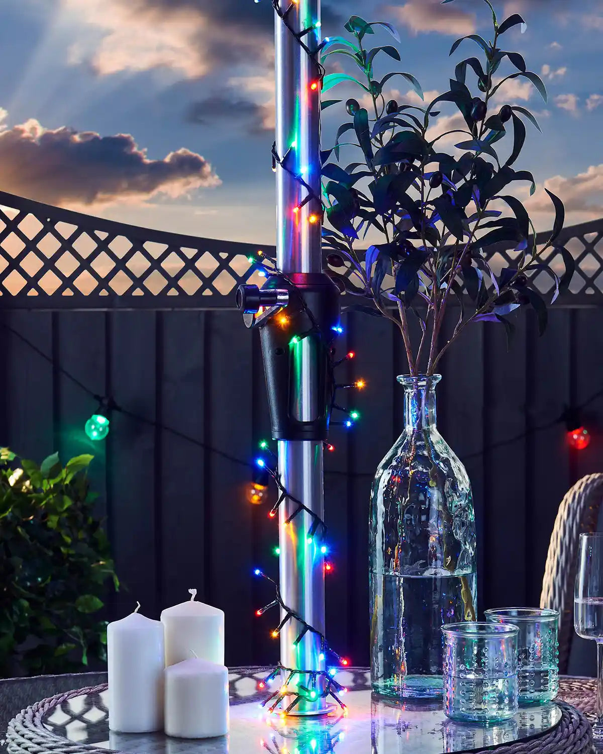 Decorative outdoor setting with a vase, candles, and multi colour string lights against a sunset sky.