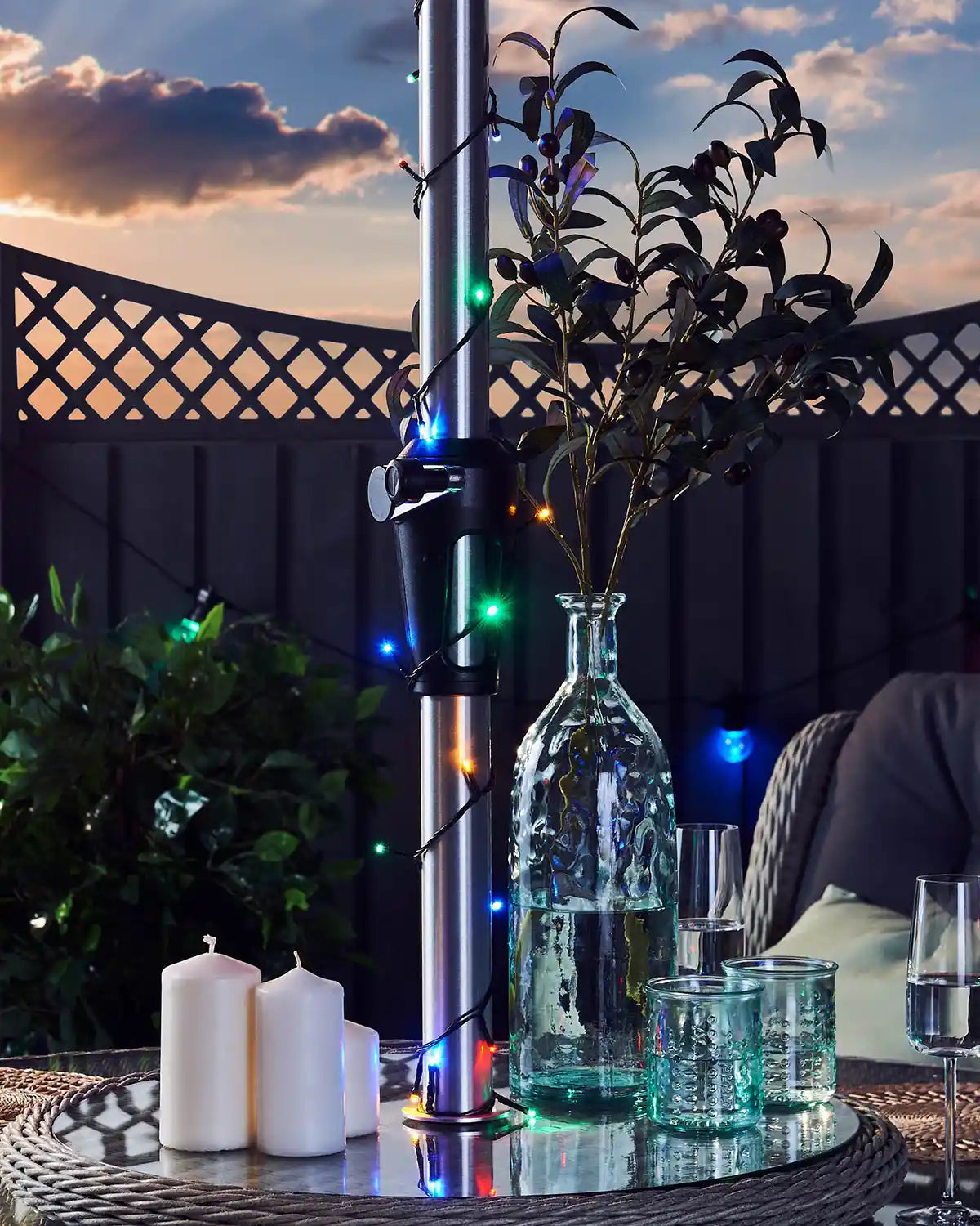 Decorative outdoor setup with a vase, multi coloured string lights, candles, and glasses on a table at sunset.