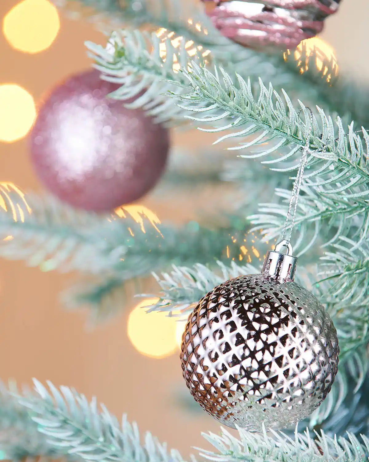 Decorative pink glitter and textured Christmas bauble on a tree branch with blurred lights in the background