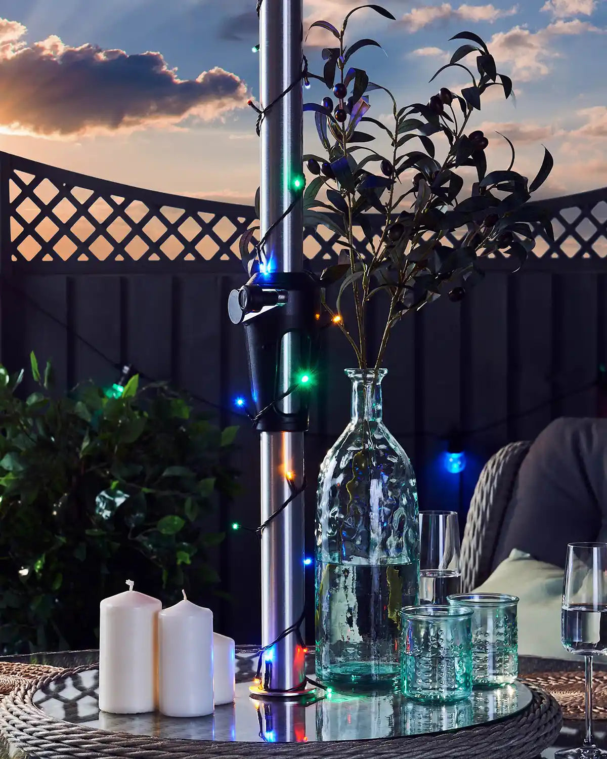 Decorative setup with a vase, multi coloured string lights wrapped around umbrella pole, candles, and glasses on a table at sunset.