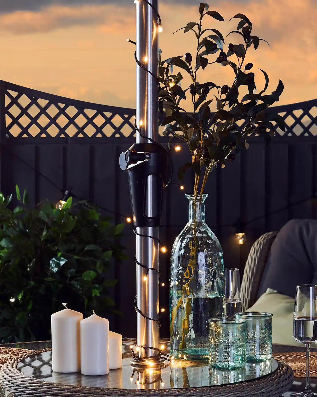 Decorative setup with candles, a vase, and warm white string lights on a table against a sunset sky.