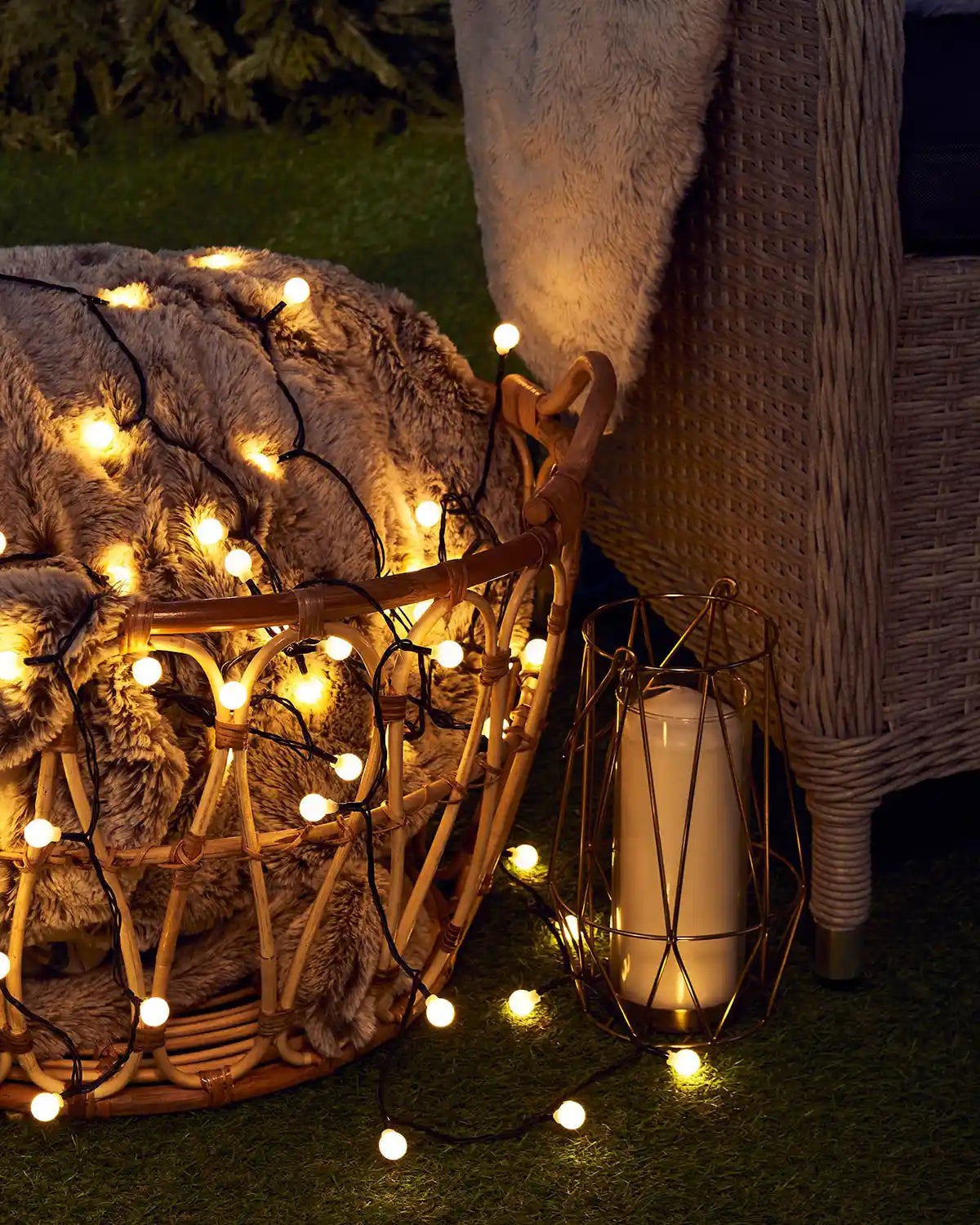 Decorative string lights on top of a blanket in a wicker basket with a candle lantern on a grassy surface.