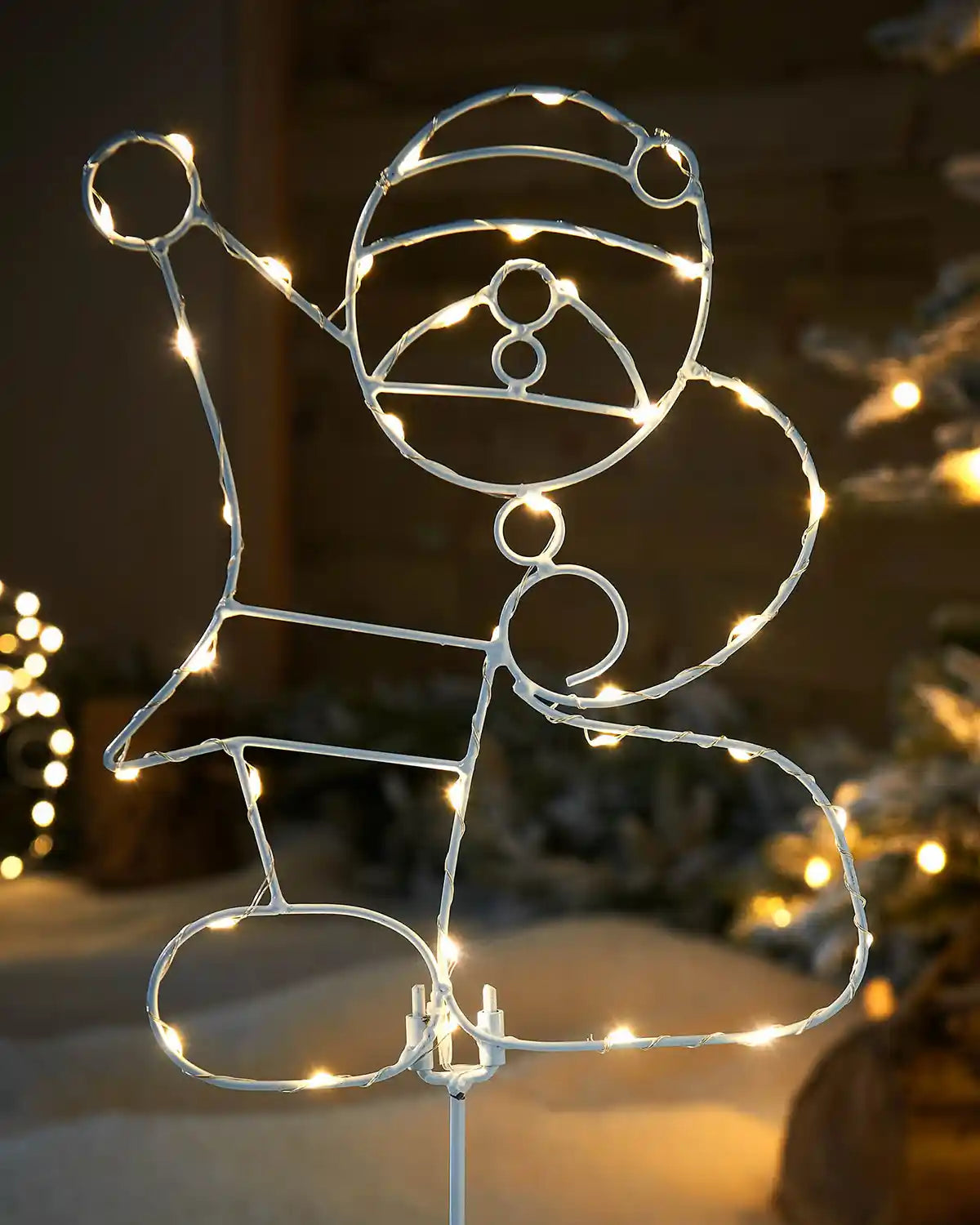 Decorative wire figure of Santa Claus with a hat and sack, illuminated by string lights against a blurred festive background.