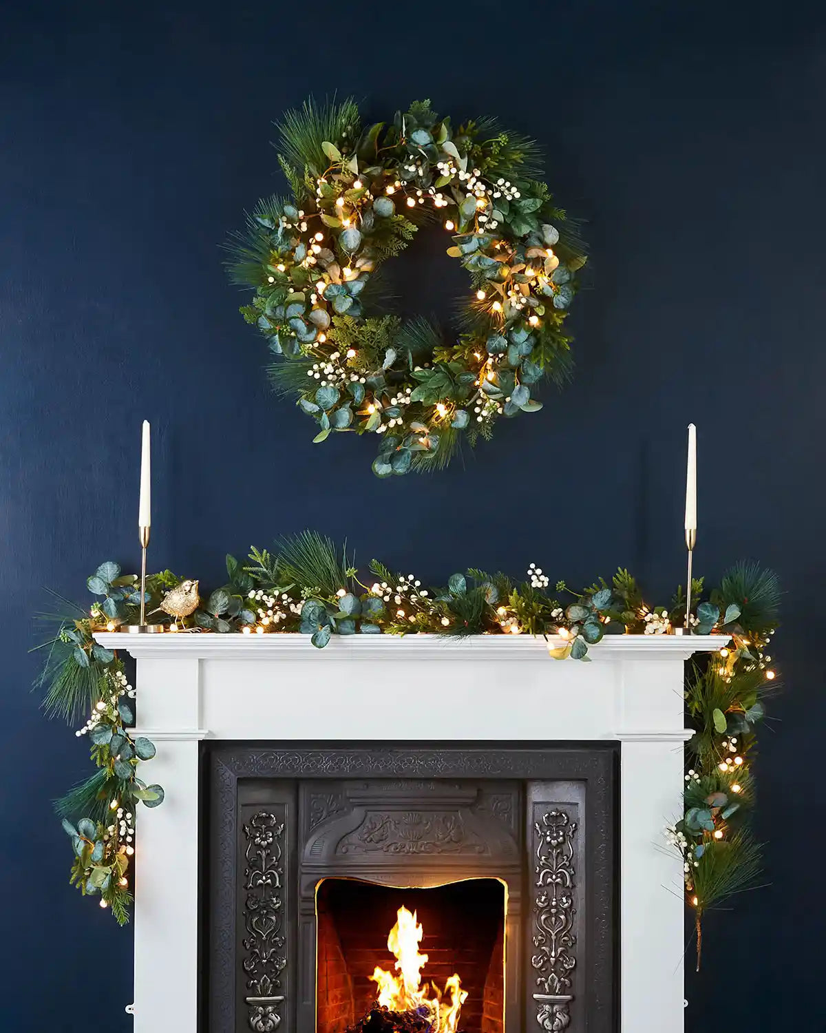 Decorative wreath with, Eucalyptus
lights and a matching garland on a fireplace against a dark blue wall.