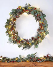 Decorative wreath with eucalyptus,  pinecones, and red berries on a white wall accompanied by a matching garland on a wooden shelf 