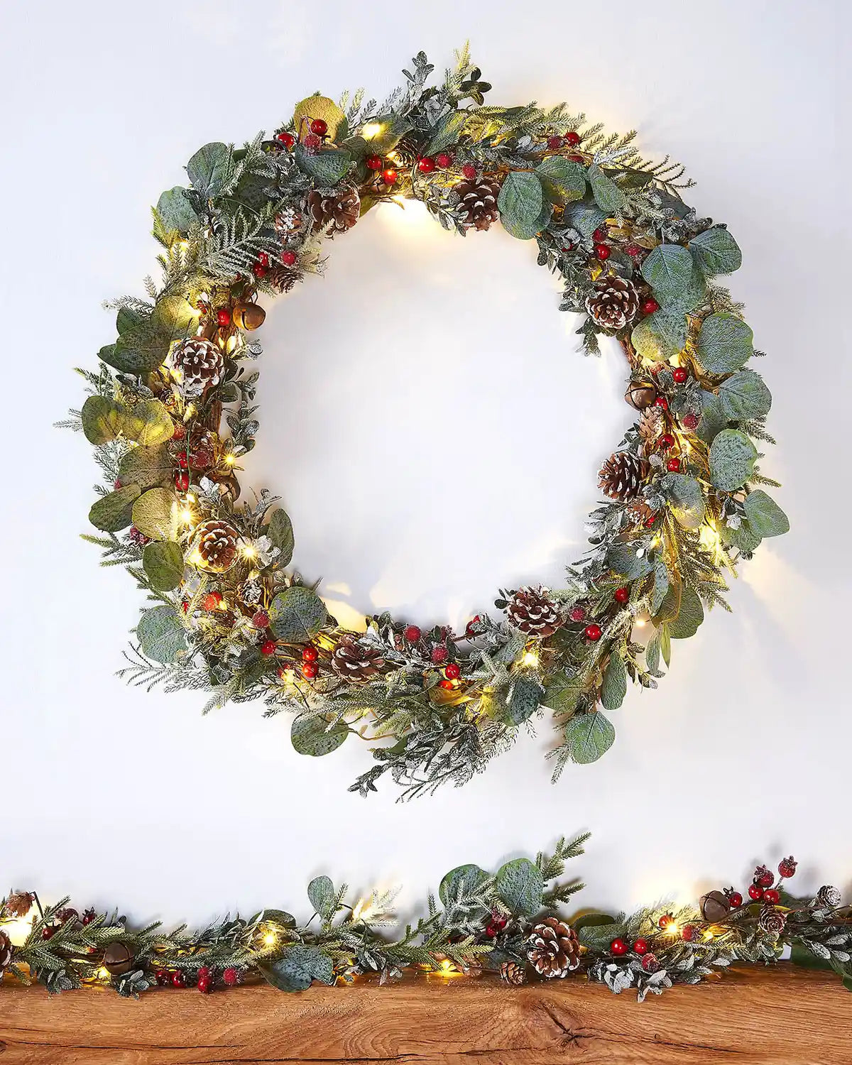Decorative wreath with eucalyptus,  pinecones, and red berries on a white wall accompanied by a matching garland on a wooden shelf 