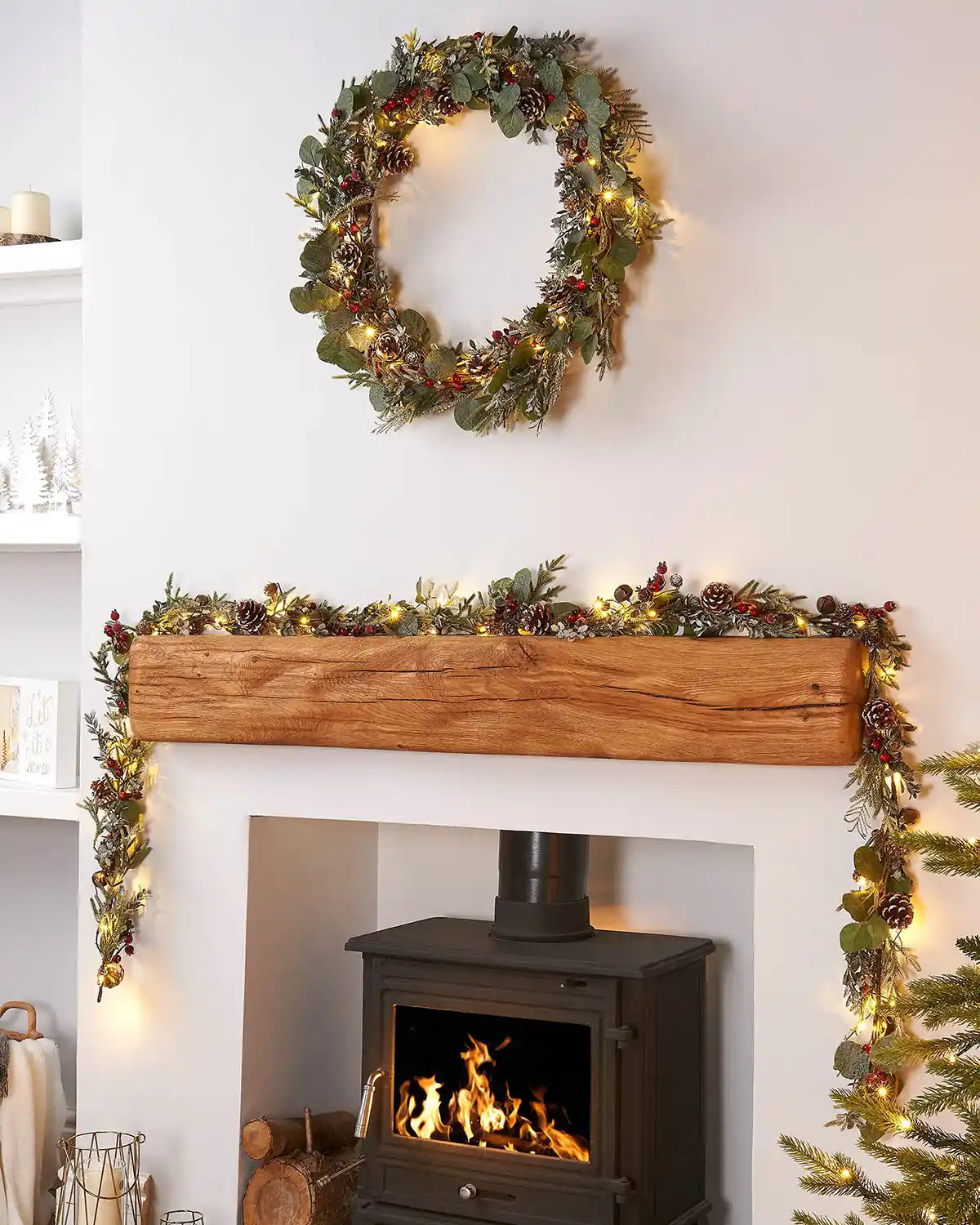 Decorative wreath with eucalyptus,  pinecones, and red berries on a white wall accompanied by a matching garland on a wooden shelf above a fireplace