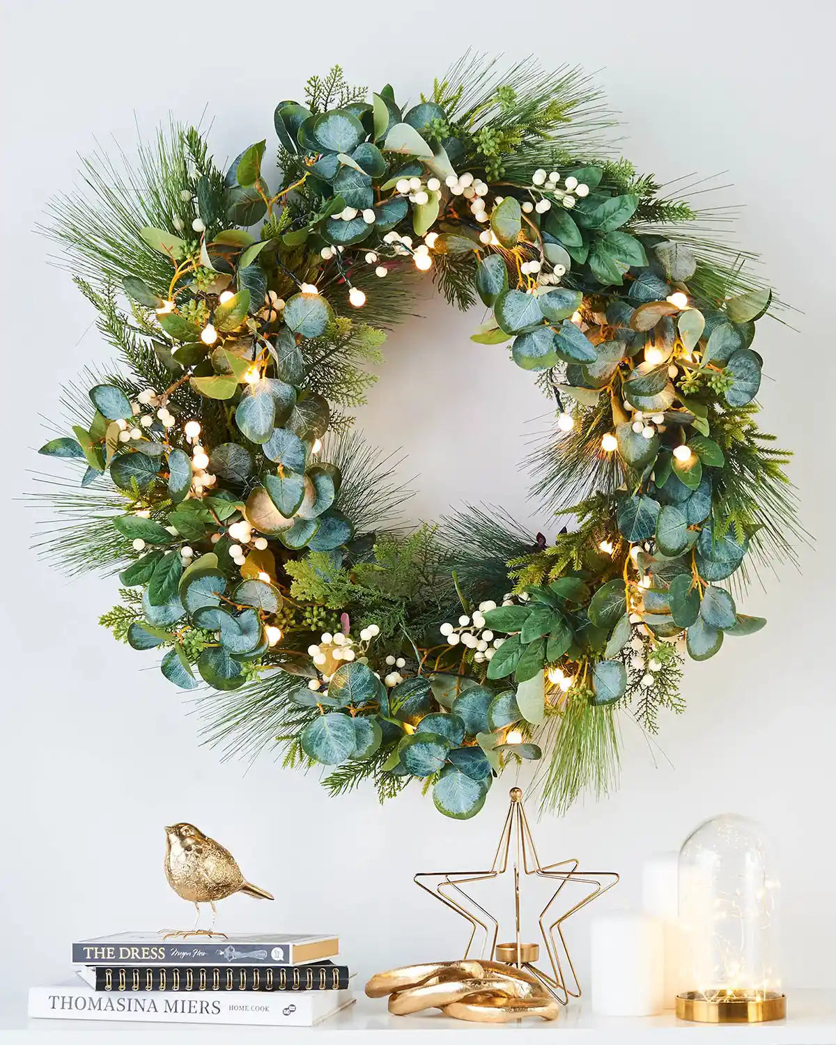 Decorative wreath with greenery, Eucalyptus and lights on a white wall with books, candles and gold ornaments 