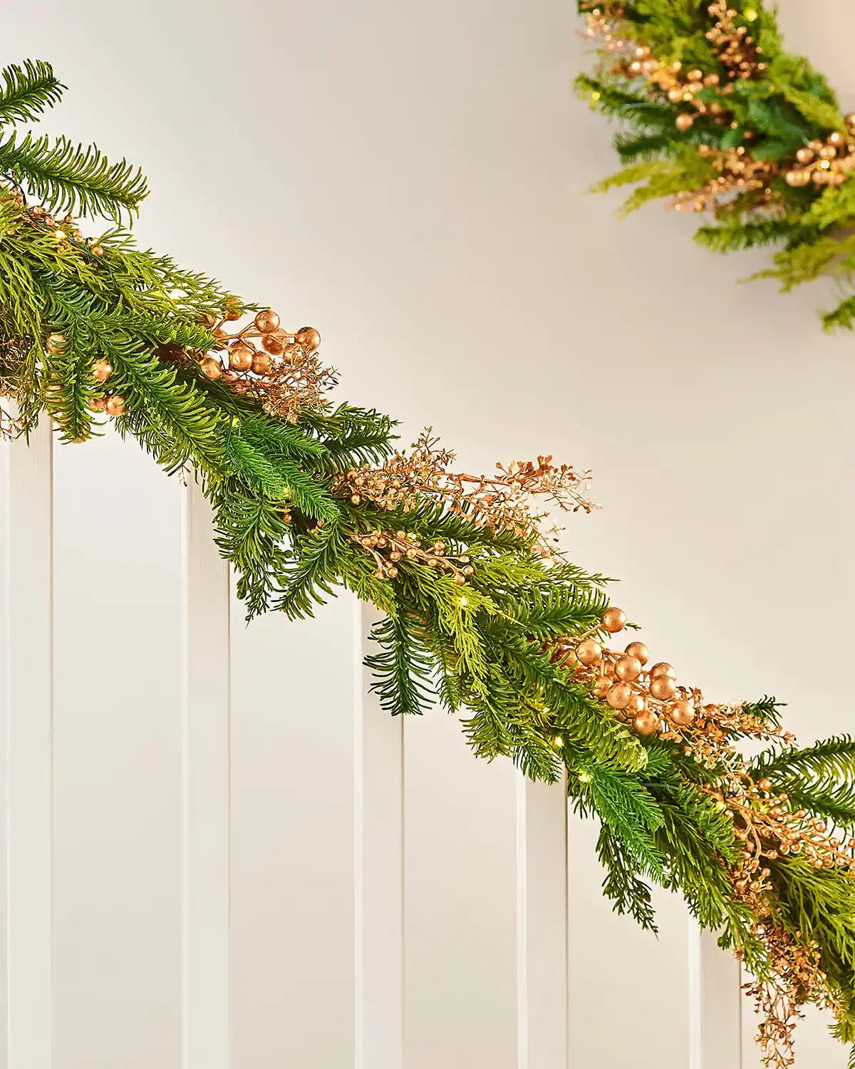Decorative wreath with lights, battery pack, greenery and small gold berries wrapped around a staircase banister on a cream background