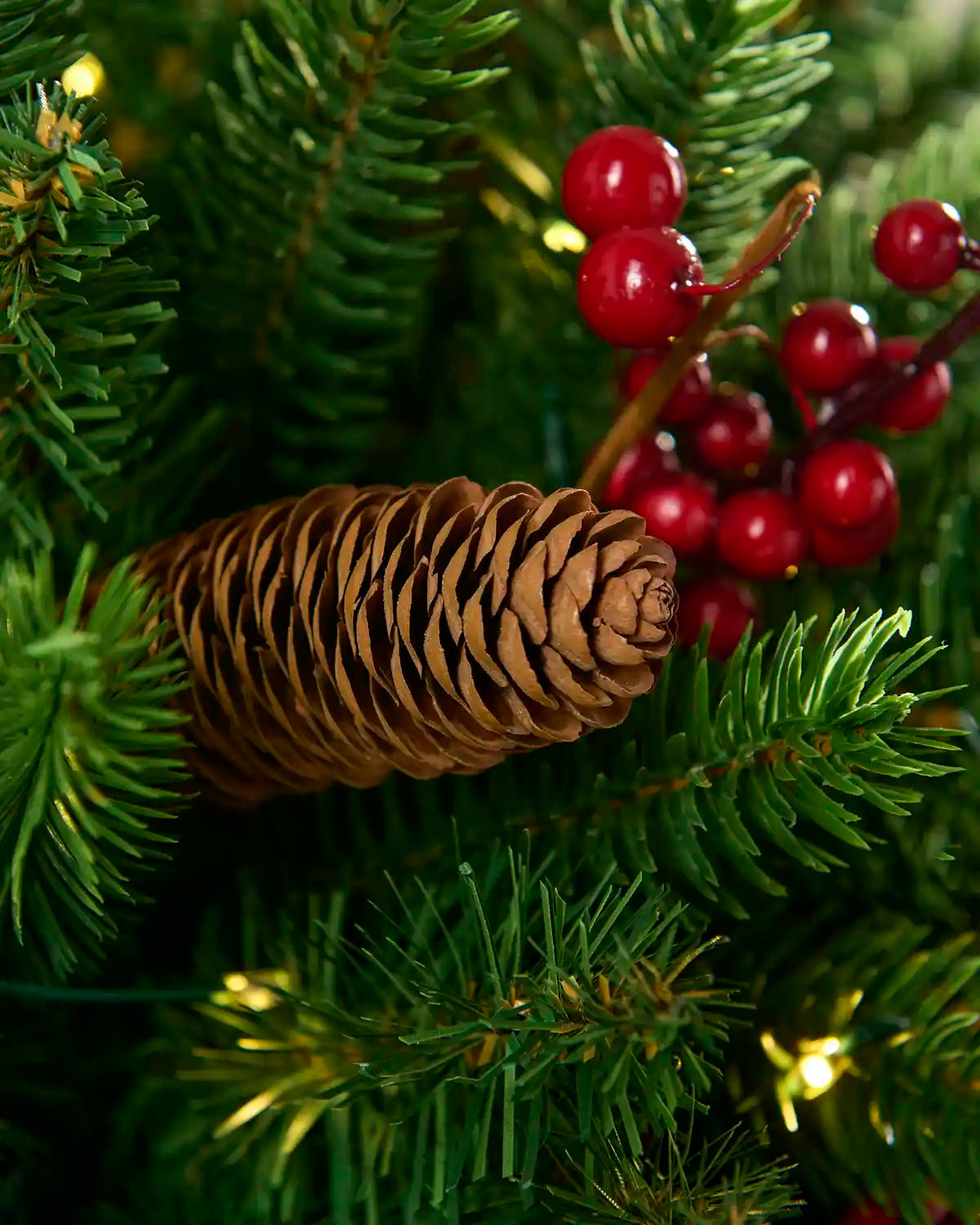 Detailed close-up of a brown pinecone nestled among green branches with red berries and warm lights