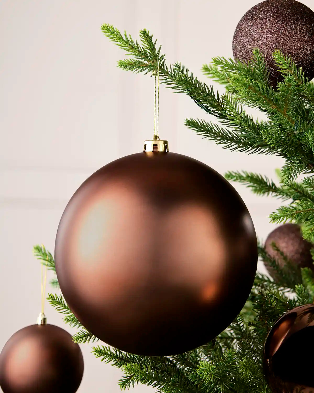 Espresso coloured oversized bauble hanging on a Christmas tree with gold string with four smaller baubles in background against a cream wall