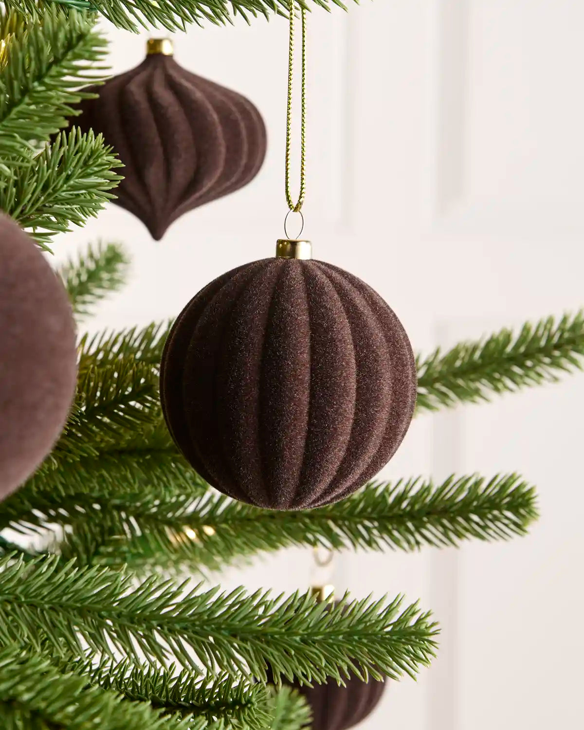 Espresso coloured velvet Christmas tree bauble hanging from hains on green pine tree branches against a light background.