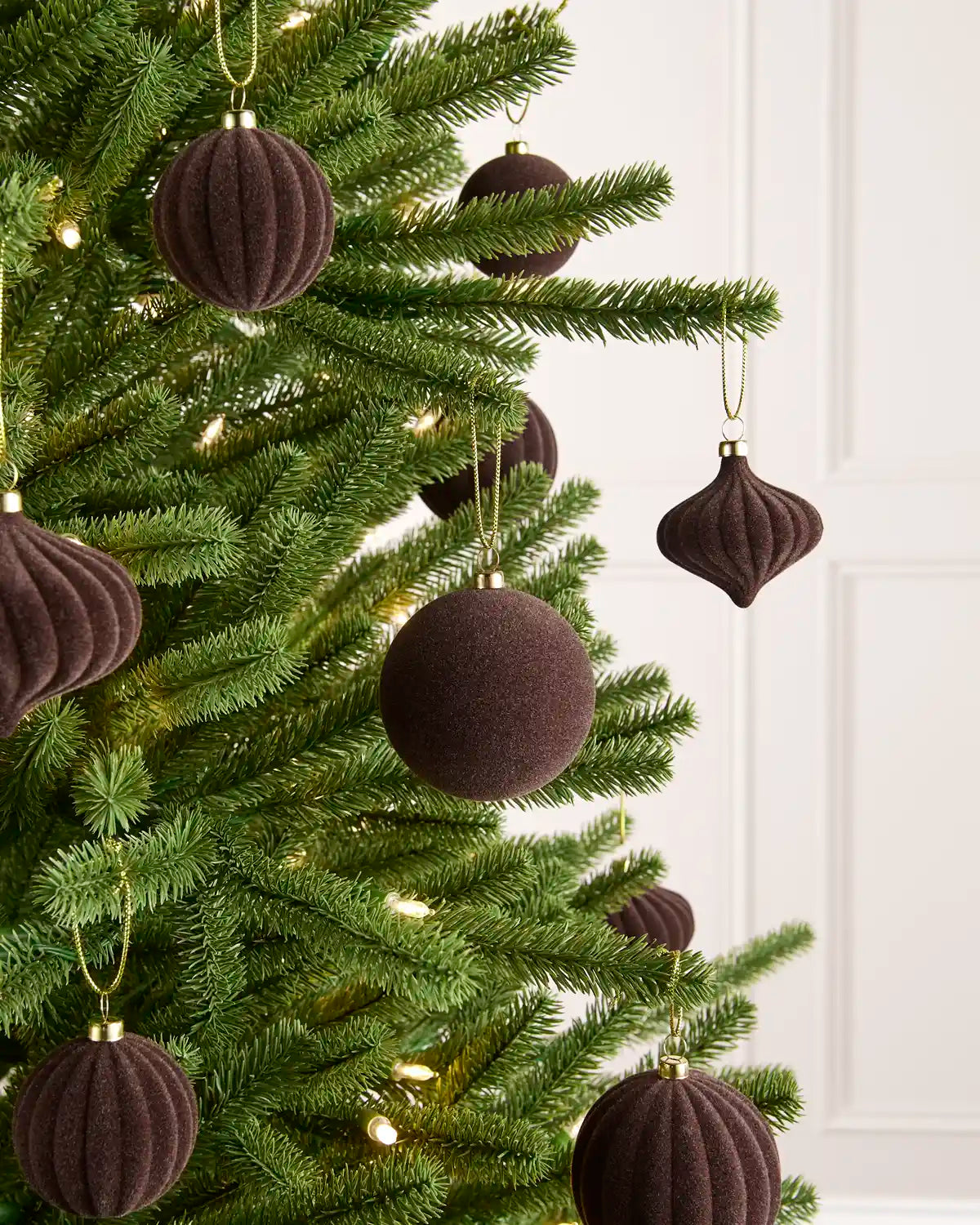 Espresso coloured velvet baubles shaped in various shapes, ribbed and smooth finishes hanging on gold string on a Christmas tree against a light background