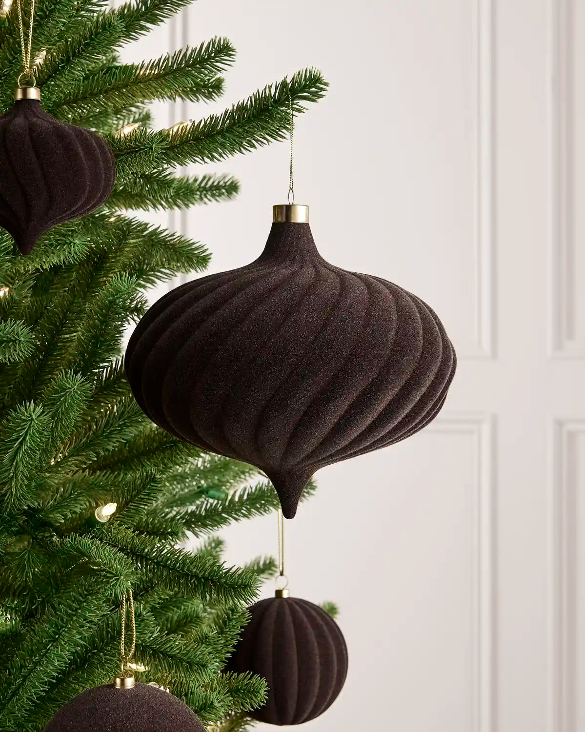 Espresso ribbed fabric Christmas tree ornaments hanging on a green Christmas tree with gold hanging loops against a white paneled wall.