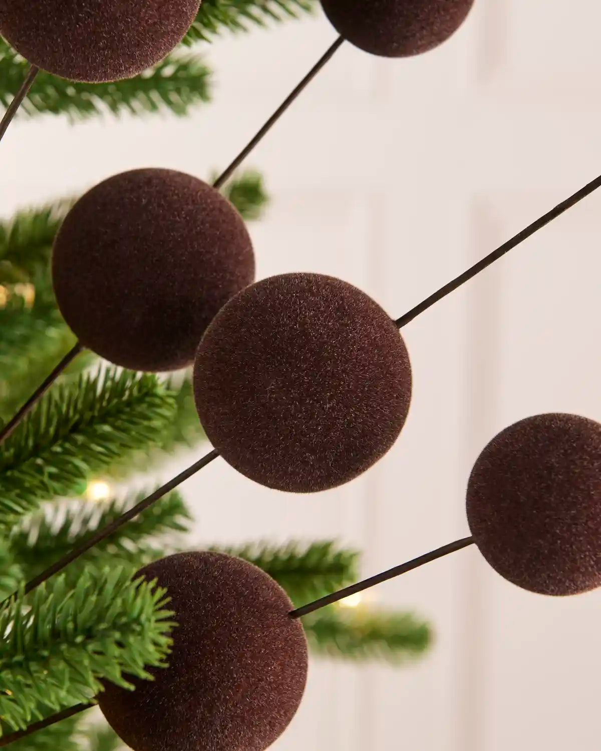 Espresso velvet bauble Christmas tree pick on thin black wire branches, shown hanging near green pine branches against a soft off-white background.