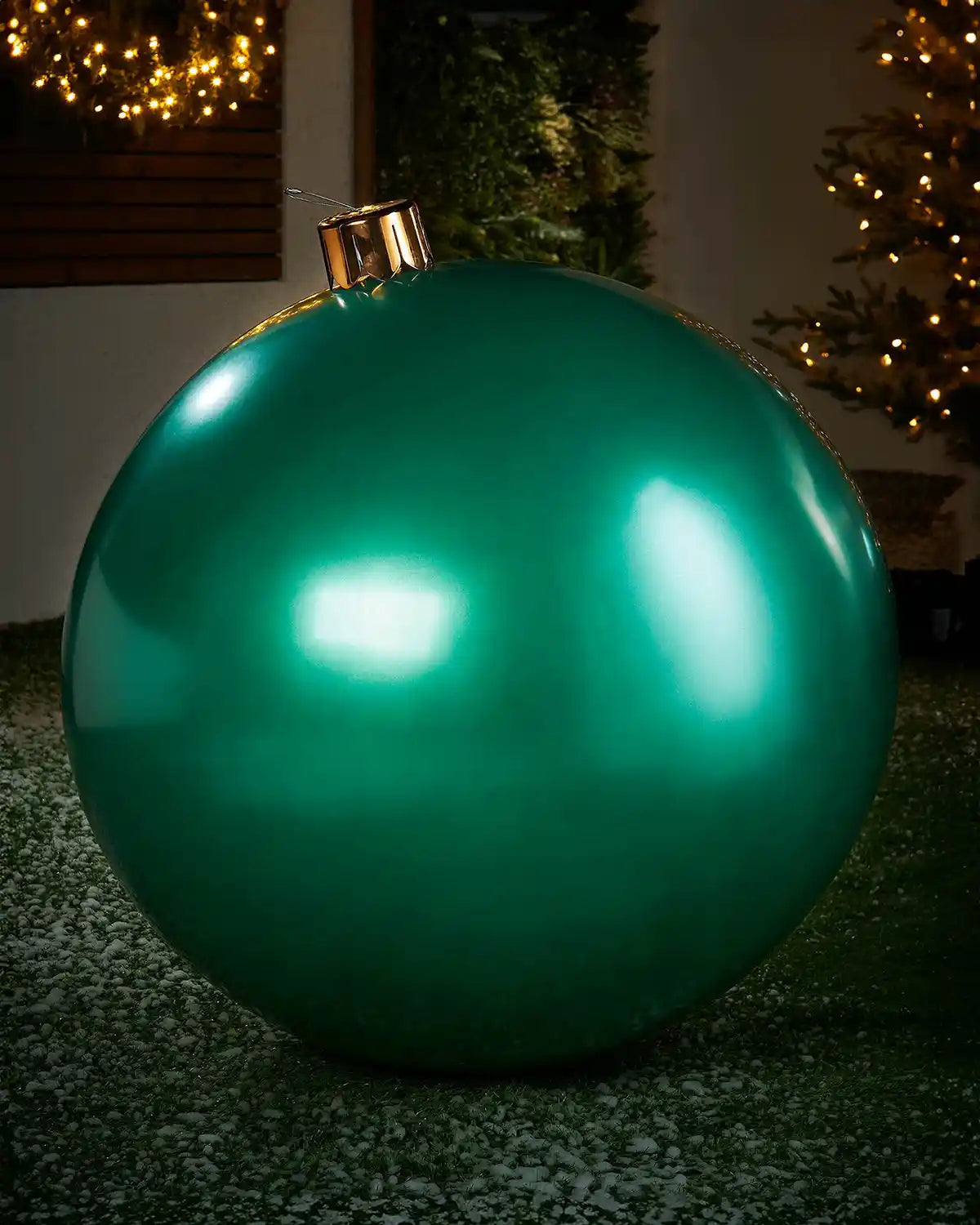 Extra large sized green inflatable bauble on a snowy garden floor with a decorated Christmas tree and wreath in background 
