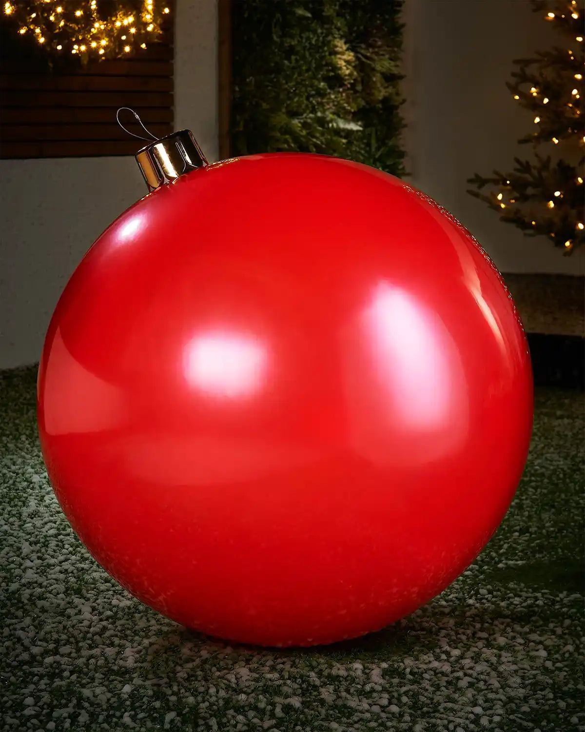 Extra large sized red inflatable bauble on a snowy garden floor with a decorated Christmas tree and wreath in background 
