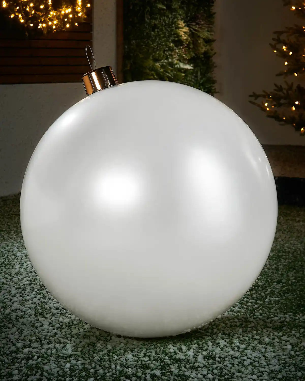 Extra large sized white inflatable bauble on a snowy garden floor with a decorated Christmas tree and wreath in background 
