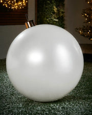 Extra large sized white inflatable bauble on a snowy garden floor with a decorated Christmas tree and wreath in background 
