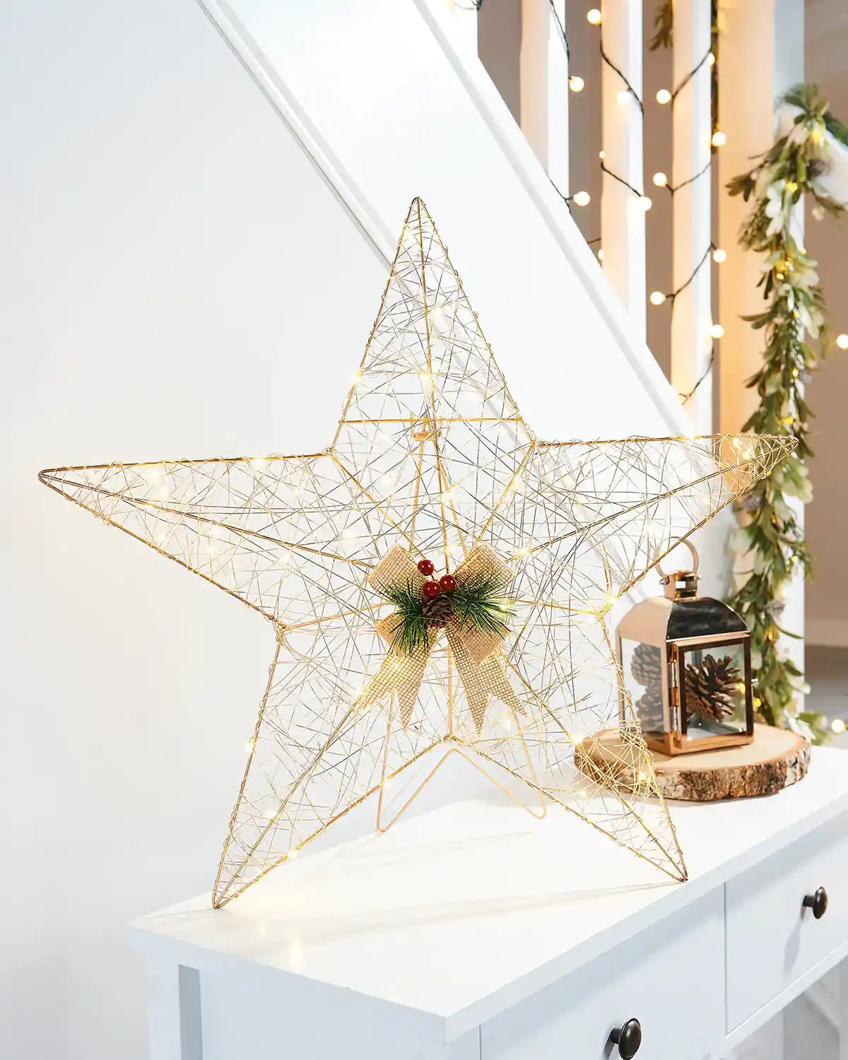 Face on view Pre-Lit 2D Metal Star, Gold, 60 cm on a White table with Pinecone lantern in front of a staircase with lights and a garland 