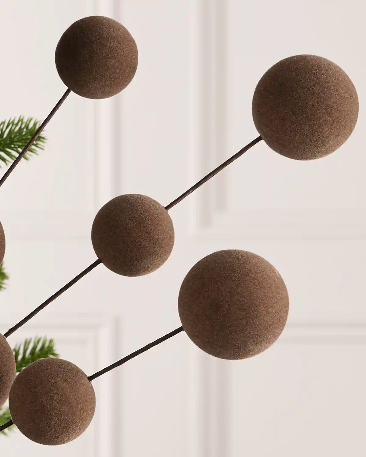 Felted brown decorative tree garland with spherical ornaments on thin black stems, shown against a soft cream paneled background with a hint of green pine foliage.