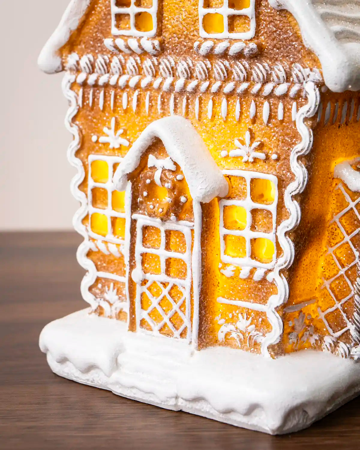 Front view of a pre-lit gingerbread house with glowing yellow windows, white icing trim, a decorated front door, and a snow-covered base