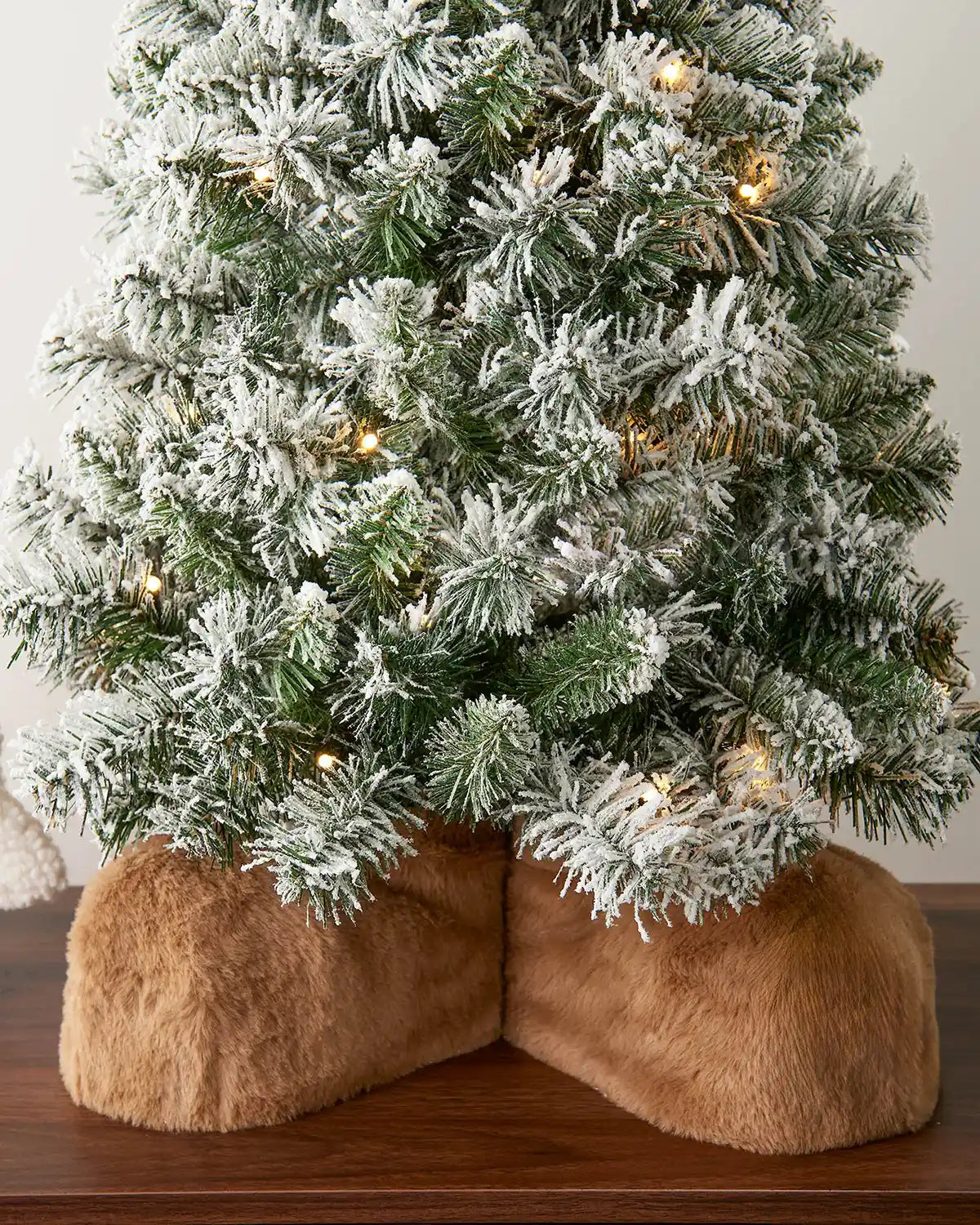 Frosted Christmas tree with lights on a wooden surface, surrounded by fluffy brown boots
