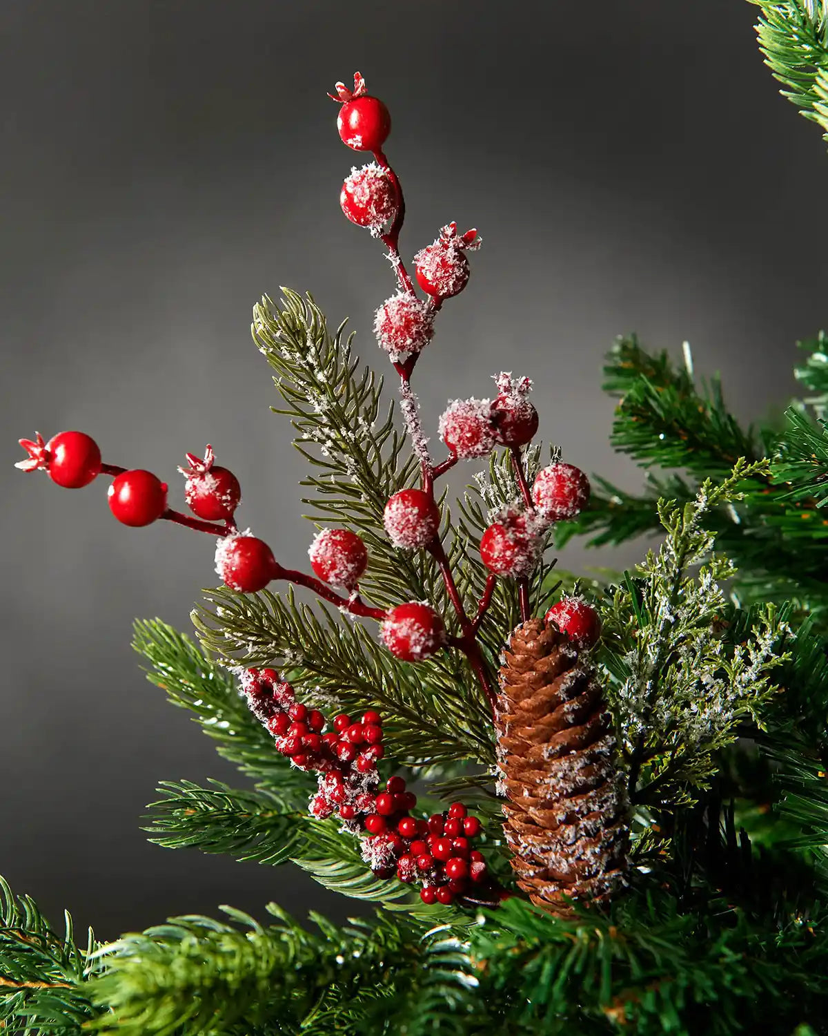 Close-up of the frosted red berries and pinecone pick attached to a Christmas tree in front of grey wall background